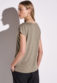 Krátký top v olivově zelené barvě s volným střihem, kulatým výstřihem a texturou slub, kombinovaný s černými kalhotami. Modelka pózuje s rukou v kapse.