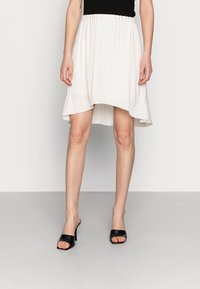 Femme portant une jupe asymétrique blanche avec une taille élastique et des escarpins noirs à bout ouvert, se tenant devant un fond gris uni.