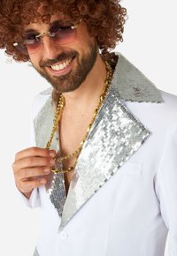 Blazer blanc avec col en sequins argentés ; collier en chaîne dorée ; cheveux bouclés ; lunettes de soleil teintées roses. Gros plan sur le haut du corps.