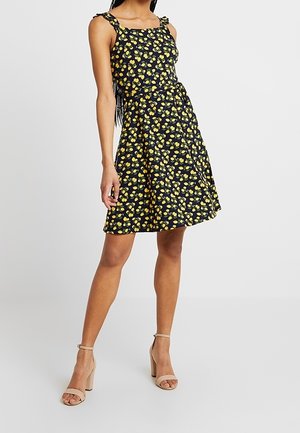 Kobieta w czarnej sukience bez rękawów z żółtym nadrukiem cytryn oraz beżowych sandałach na wysokim obcasie, stojąca na jednolitym tle.