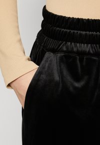 Sorte fløyelsbukser med elastisk midje vist sammen med en ribbet, beige langermet topp, som fremhever lomme detaljen.