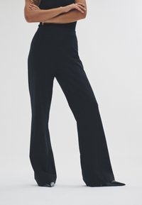 Højttaljede sorte bukser med viddeben, glat stof og diskrete lodrette center-søm detaljer. Afsluttet med skræddersyede benåbninger.