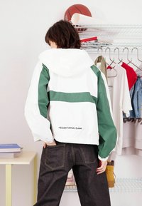 Biała i zielona kurtka przeciwdeszczowa z kapturem, z poziomym zielonym paskiem i elastycznymi mankietami. Noszona z ciemnymi dżinsami, prezentująca casualowy styl.