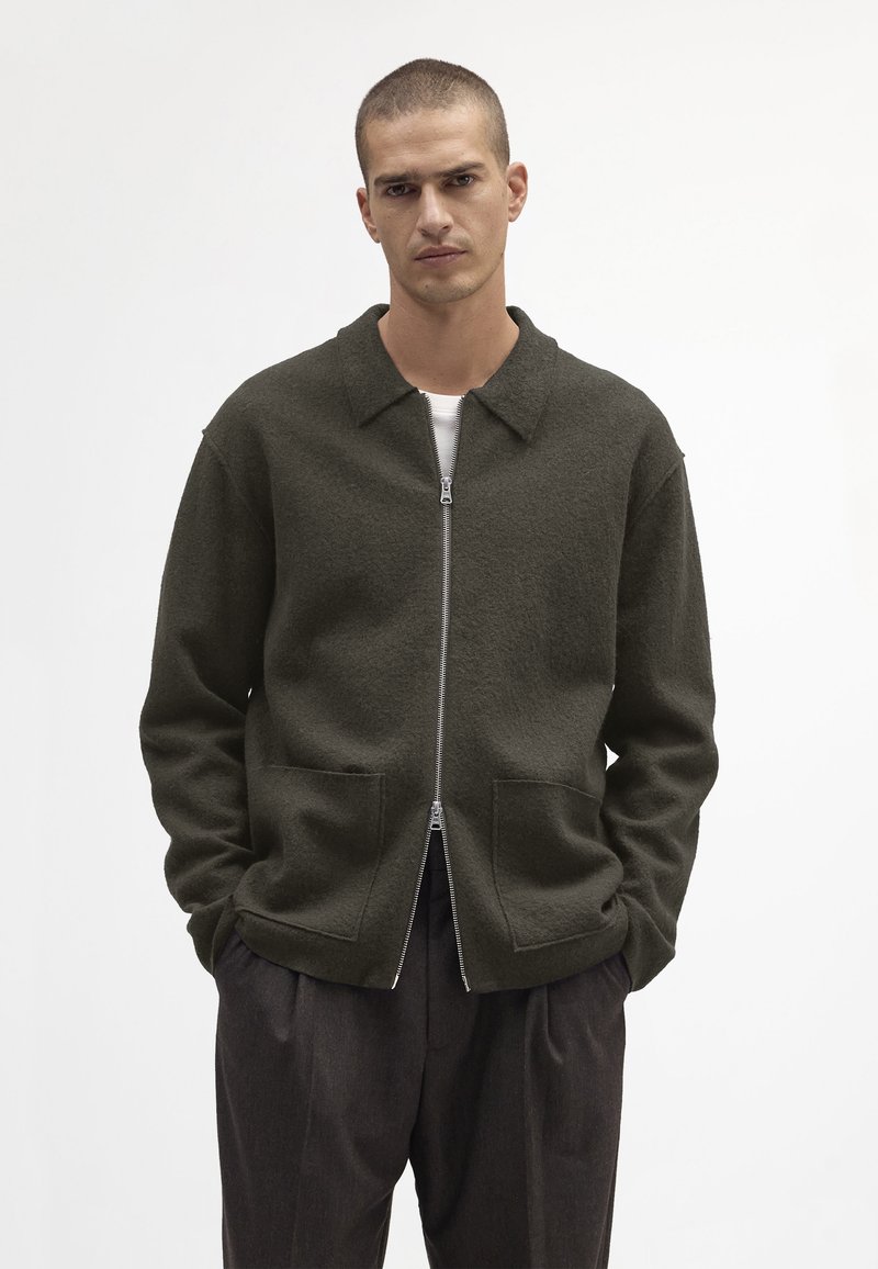 Dunkelgrüne Fleecejacke mit Kragen, Reißverschluss und zwei Fronttaschen. Kombiniert mit dunklen Hosen und einer weißen Innenschicht.