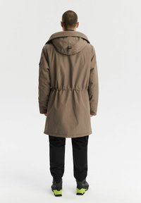 Brązowy parka z kapturem i ściągnięciem w talii, o gładkiej teksturze. Nosi ją z czarnymi spodniami i sportowymi butami z zielonymi akcentami.