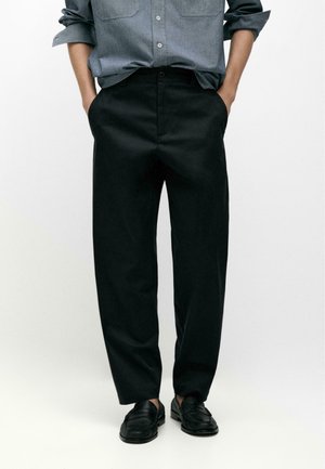 Persona che indossa pantaloni neri a gamba dritta, mocassini neri e una camicia grigia a maniche lunghe con le maniche arrotolate, con le mani in tasca.