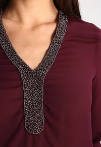 Blouse bordeaux avec décolleté en V et détails décoratifs en perles argentées. Le tissu a une texture lisse et une coupe structurée.