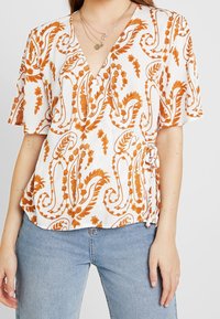 Blouse blanche avec un motif paisley orange, décolleté en V, manches courtes et détail de nouage sur le côté. Tissu doux et texturé avec une coupe décontractée.