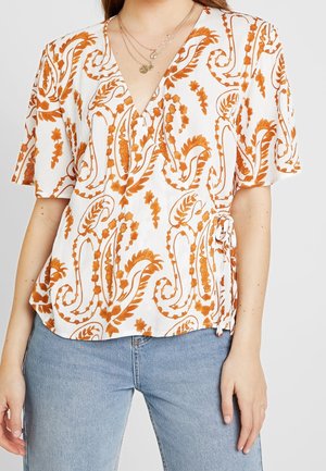 Blouse blanche avec un motif paisley orange, décolleté en V, manches courtes et détail de nouage sur le côté. Tissu doux et texturé avec une coupe décontractée.