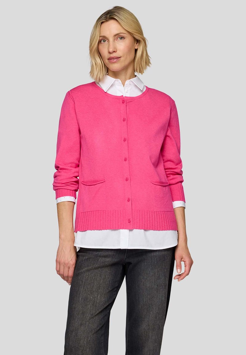 Pinkfarbener Cardigan mit rundem Ausschnitt, Knopfleiste vorne, zwei Fronttaschen, geripptem Saum und langen Ärmeln, getragen über einem weißen Hemd mit Kragen.
