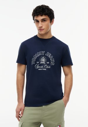 Young man with short curly hair wearing navy blue "Tommy Jeans Sports Club New York" T-shirt and light green cargo pants, standing with hand in pocket.