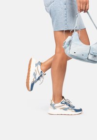 Zapatillas deportivas en azul y blanco con parte superior de malla, detalles texturizados, sobreposiciones estampadas y una suela de goma gruesa. Llevadas con una falda de mezclilla.