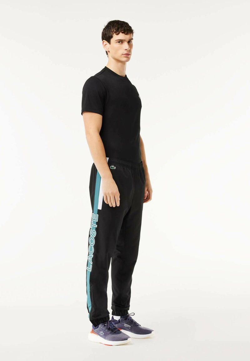 Chemise noire à manches courtes et pantalon de survêtement noir avec des accents turquoise et le logo "LACOSTE". Le modèle porte des chaussures de sport navy avec des détails orange.