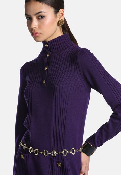 Femme portant un pull violet côtelé à col montant avec des boutons dorés, une ceinture chaîne dorée, de grandes boucles d'oreilles dorées et un bracelet manchette noir et or.