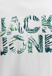Bela bombažna majica s kratkimi rokavi, na kateri je velika napis "JACK & JONES" v vzorcu zelenih palmih listov. Mehka tekstura, priložnostna zasnova.