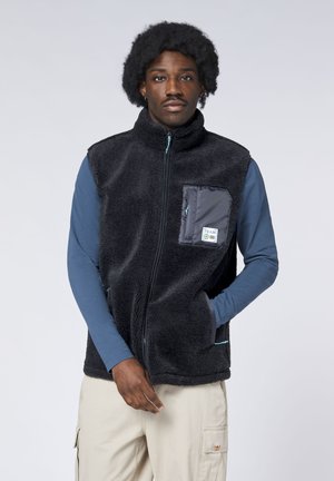 Man wearing black fleece vest over blue long-sleeve shirt and beige cargo pants, standing against plain light background.