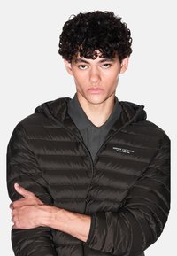 Giacca imbottita nera con cappuccio, texture trapuntata, cerniera frontale e logo sul petto. Modello con le braccia incrociate, indossa una polo grigia sotto.