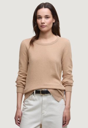 Frau mit langen braunen Haaren trägt einen beigen Pullover und weiße Hose mit schwarzem Gürtel, steht vor einem schlichten, hellen Hintergrund.