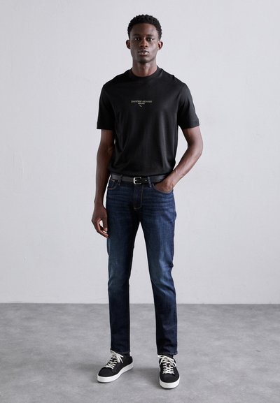 T-shirt noir à manches courtes avec logo, porté avec un jean bleu foncé et des baskets noires. Le modèle se tient devant un fond neutre.