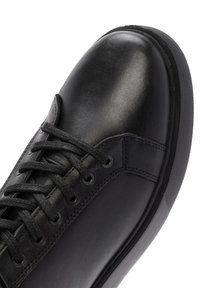 Black leather sneaker with round toe, flat laces, metal eyelets, textured upper, and contrasting rubber sole. Features no visible logos.