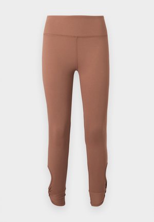 Hnedé legíny s vysokým pásom a nariasenými detailmi pri členku, zobrazené na jednoduchom svetlom pozadí.