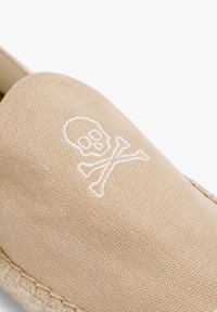 Beige canvas instappers met een geborduurd doodskop en gekruiste botten ontwerp, voorzien van een geweven jute zool en een gladde textuur.