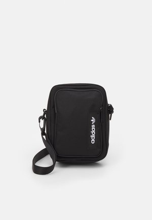 Petit sac bandoulière noir Adidas avec poche zippée à l'avant et bandoulière réglable sur fond blanc.
