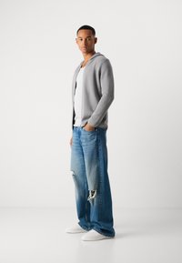 Lys grå cardigan med hette, båret over en hvit t-skjorte, kombinert med slitte blå jeans og hvite joggesko. Slim fit og avslappet stil.