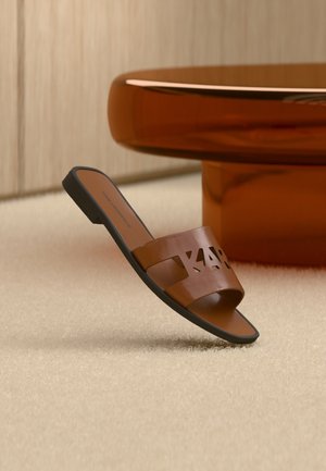 Καφέ δερμάτινο σανδάλι σλάιντ με γράμματα κομμένα σε μπεζ χαλί δίπλα σε γυαλιστερή στρογγυλή βάση τραπεζιού κεχριμπαρένιου χρώματος.