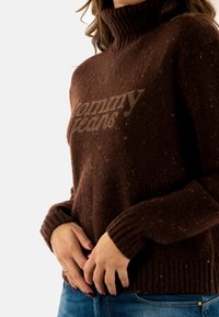 Maglione lavorato a maglia marrone con colletto alto, presenta un logo in rilievo "Tommy Jeans" in beige e spruzzi multicolori su tutta la stoffa.