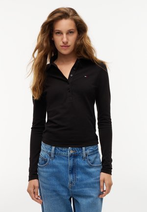 Kvinde med langt bølget hår iført en sort langærmet polo og lyseblå jeans, står foran en hvid baggrund.