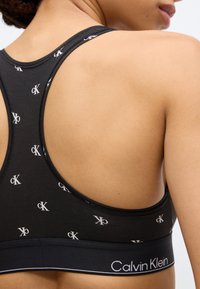Μαύρο αθλητικό σουτιέν με επαναλαμβανόμενο λευκό μονόγραμμα "ck", σχέδιο racerback και μαύρη σταθερή ταινία με το κείμενο "Calvin Klein" σε λευκό.
