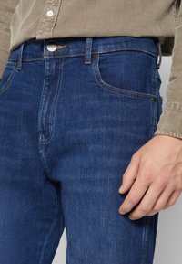 Jeans de denim azul oscuro con un clásico diseño de cinco bolsillos, costuras en contraste de color naranja y un cierre de botón en la cintura.