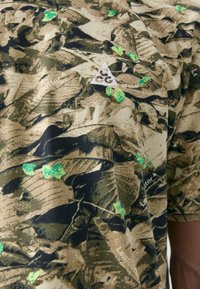 Short-sleeve shirt with a green and brown leaf camouflage pattern, featuring a white triangular logo on the upper left. Cotton material.