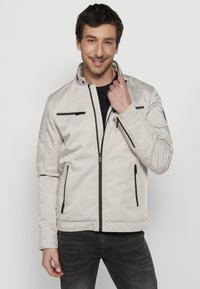 Svetlo béžová bomber bunda s čiernymi zipsovými vreckami, predným zipsom a stojacím golierom. Hladká textura s štruktúrovaným strihom.