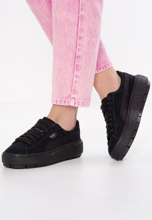Beine in pinken Jeans und schwarzen Puma Plateau-Sneakern stehen auf weißem Untergrund, ein Fuß leicht angehoben.