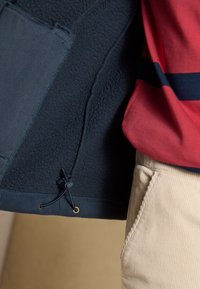 Kordelzug-Detail am inneren Saum einer dunklen Fleecejacke, getragen mit beige Cordhosen und einem rot-navy gestreiften Oberteil.