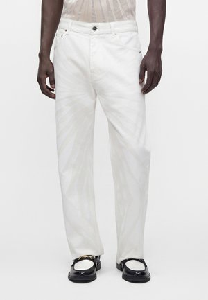Homme portant un pantalon blanc droit et des mocassins noir et blanc, debout devant un fond uni.