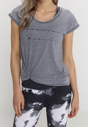 Kobieta ubrana w szarą, zawiązaną koszulkę z napisem „Venice Beach” oraz czarno-białe legginsy tie-dye, ukazująca tułów i górną część nóg.