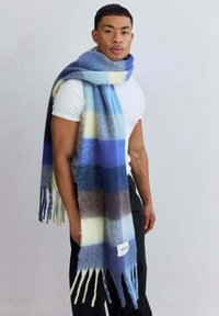 Blauwe en gele gestreepte sjaal van zacht materiaal, met franjes aan de uiteinden en een label met de tekst "ARCTIC 20." Om de nek van een model gedrapeerd.