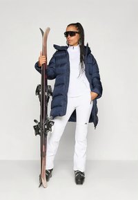 Pufferjakke i marineblå med en teksturert overflate, kombinert med hvite skibukser med horisontale striper; modellen holder ski og har på seg solbriller.