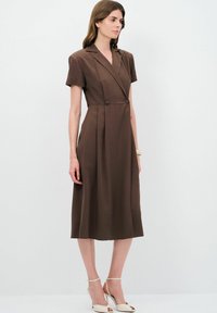 Robe marron, longueur genou, avec des manches courtes, décolleté en V, devant double boutonnage et jupe évasée. Tissu lisse avec un subtil détail de drapée.