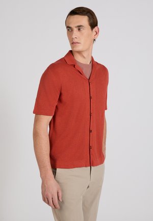 Mann trägt rostfarbenes kurzärmliges Hemd über einem braunen Shirt, mit beigen Hosen, blickt seitlich vor einem schlichten Hintergrund.