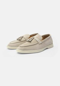 Mocassini in suede beige con dettaglio a nappina, punta arrotondata, suola bianca a contrasto e dettagli cuciti lungo il bordo superiore.