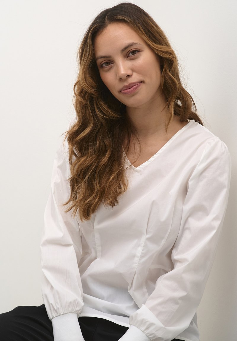 Weiße Bluse mit langen Ärmeln und V-Ausschnitt, mit Puffärmeln und glatter Textur. Sichtbare Manschetten, kombiniert mit schwarzen Hosen.