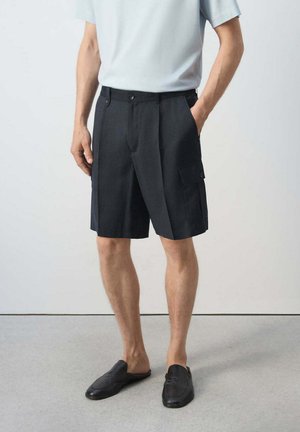 Homme portant un short cargo noir avec poches boutonnées, une chemise blanche à manches courtes et des chaussures noires sans lacets, debout avec une main dans la poche.