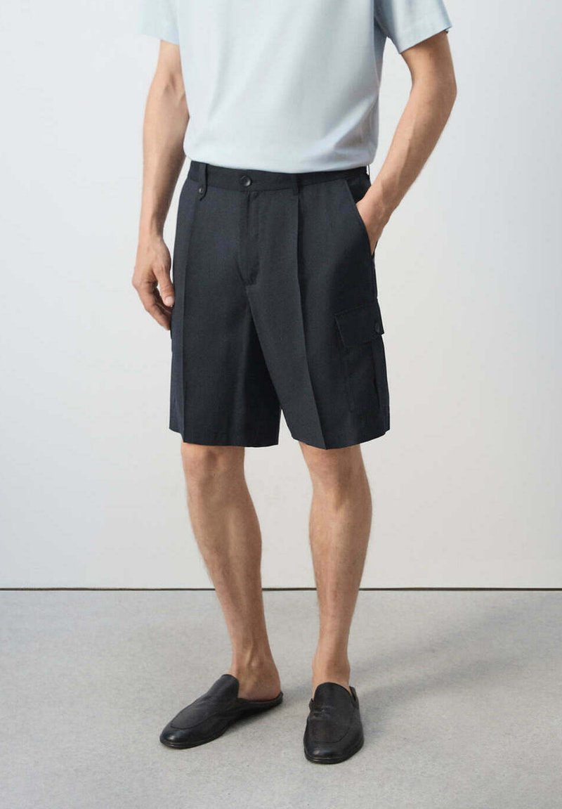 Homme portant un short cargo noir avec poches boutonnées, une chemise blanche à manches courtes et des chaussures noires sans lacets, debout avec une main dans la poche.