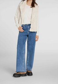 Krämfärgad väst med fårskinnstextur över en vit långärmad topp, kombinerad med blå vida jeans och bruna-svarta platåskor.