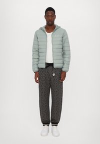 Lys grøn pufferjakke med hætte, sorte mønstrede joggers og hvide sneakers. Jakken har lodret syning og logo.
