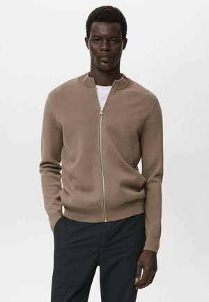 Man wearing beige zip-up sweater over white shirt and dark pants, standing with one hand in pocket against plain background.